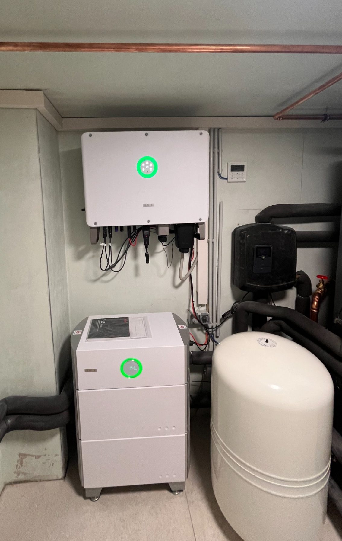 SAJSAJ H2-25K-T3 Hybrid – Wechselrichter 25 kW Zwei Geräte an einer Wand: oberhalb ein Steuergerät, darunter eine Heizungsanlage mit Pumpe.